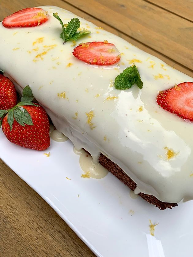Strawberry Bread With or Without Gluten and Dairy with white icing, decorated with strawberries, mint, lemon peel, on a white plate with whole strawberries. Wooden table in background.
