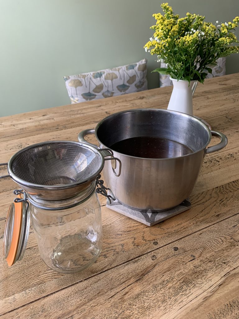 Bone broth in a large saucepan and glass jar with funnel and sieve in foreground