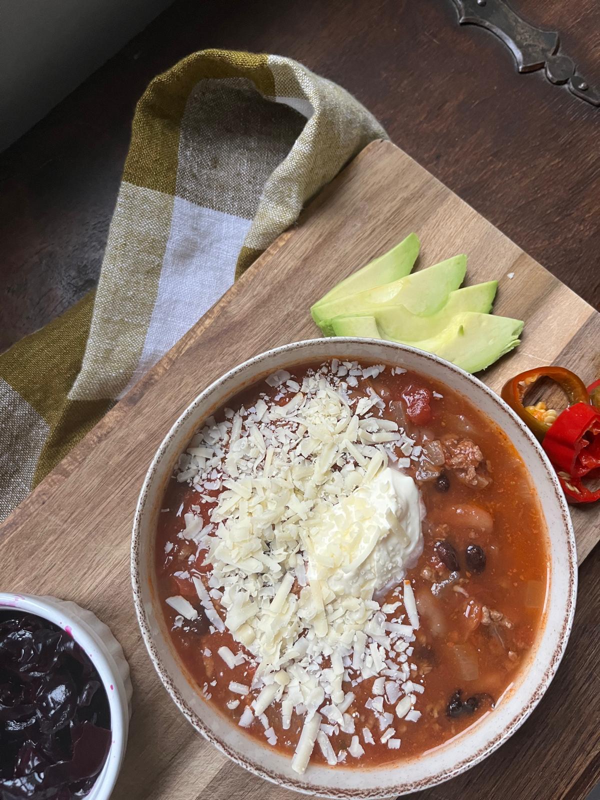 bowl of easy ground beef chili topped with cream and cheese and served on a board with red sauerkraut, fermented chili peppers and avocado slices